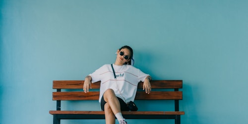 Young adult woman sat on bench in baggy t-shirt & sunglasses against blue background